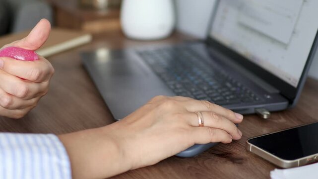 Woman squeezing one hand pink squishy stress toy while other work on laptop using computer mouse in 4k footage. Concept of multitasking, work stress relief and tactile coping during digital tasks.