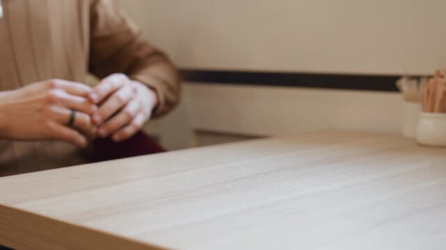 Close up of man hands resting on wooden table fingers interlaced and relaxed. Man wears brown long sleeve shirt with zipper detail soft lighting creates warm ambiance. Background includes