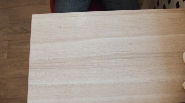 Close up of person hands resting on wooden table fingers interlaced and gently moving. person wears brown long sleeved shirt with subtle vertical stripes setting includes small objects