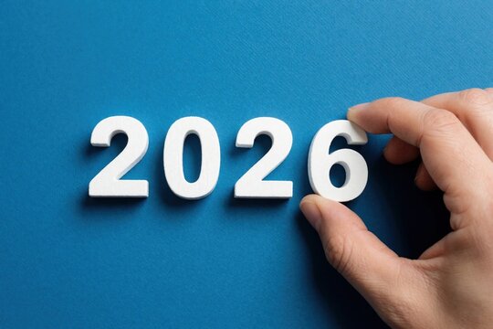 Human hand places white number 6 to complete 2026 on blue background. Person finishing year digits for new year planning. Concept of goal achievement, future strategy, time transition. Minimalist