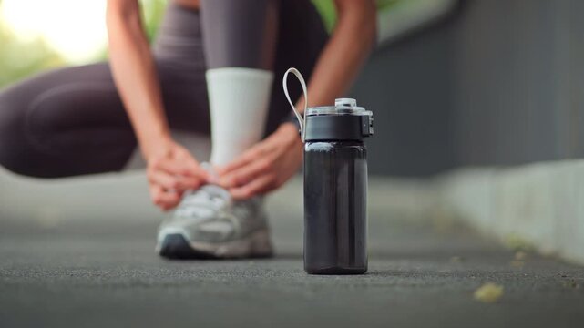 Close up of reusable water bottle in foreground while young Asian woman tying running shoes before outdoor workout, preparing for exercise, hydration, fitness training and healthy active lifestyle con