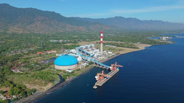 Aerial view of Celukan Bawang Steam Power Plant with its distinctive blue storage dome, tall chimney, and jetty on the coastline of Buleleng, Bali, Indonesia.