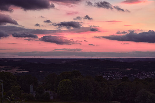 Soft pink sunset sky casts warm light over rolling hills, a small village and distant wind turbines, creating a peaceful, scenic evening landscape filled with color and atmosphere.