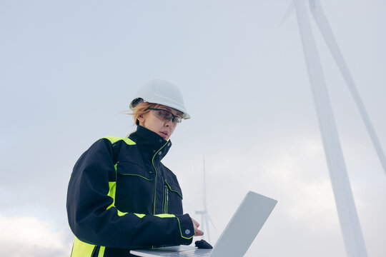 Woman engineer in hard hat and safety glasses using laptop near windmills