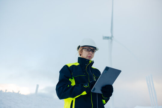 Woman engineer in safety gear checks tablet near wind turbine in snowy landscape