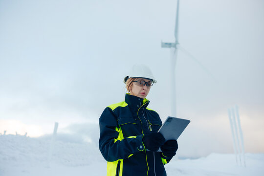 Woman engineer in safety gear checks tablet near wind turbines in snowy landscape