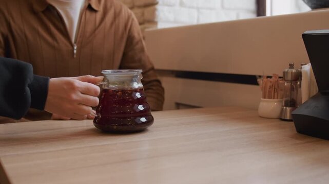 Close up of man in brown jacket sitting at wooden table with condiment set. Another man places glass coffee pot and cup on same tabletop then moves away. setting is indoor cafe or restaurant