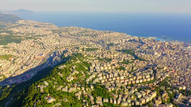 Genoa, Italy. Aerial panorama over the historic Porto Antico and dense terracotta rooftops, with cargo ships and cranes lining the deep blue Ligurian Sea under bright summer light. Drone footage