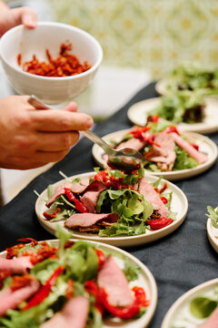 Slices of roast beef atop fresh greens and red peppers, plated for a family-style dinner. A bowl of spices or toppings is being sprinkled, creating a warm, inviting meal scene.