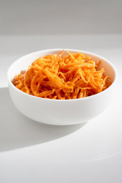 bowl of finely shredded orange carrots sits on plain white surface, highlighting fresh, healthy ingredients. Perfect for recipe, vegan, or minimalist food photography emphasizing color and simplicity.