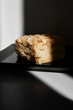 Single slice of layered almond napoleon cake rests on a sleek black plate, illuminated by dramatic light. Rich textures, golden crumbs, and rustic charm evoke premium bakery desserts.