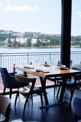 Fototapeta premium Sunlit modern restaurant dining table set with chairs and glassware by a large window overlooking a calm river and distant town, evoking relaxed waterfront dining, leisure, and elegant hospitality.