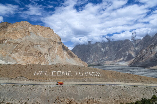 View of the Welcome to Pasu sign on a rugged hillside with a truck on the road and the dramatic Passu Cones mountain range in Hunza Nagar, Gilgit Baltistan, Pakistan.