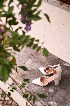 White bridal heels rest on weathered stone steps surrounded by soft green leaves and scattered petals, evoking elegant wedding preparation, romance and minimalist outdoor fashion detail.
