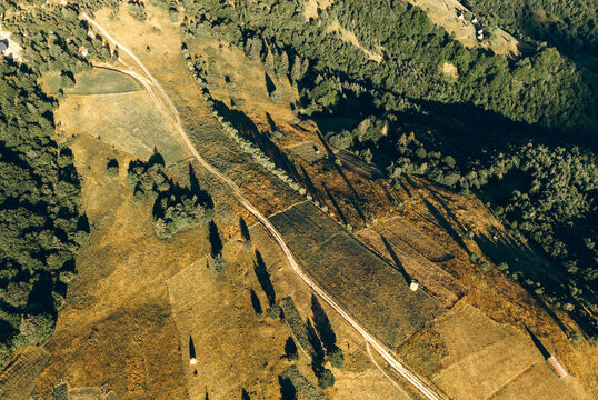 Aerial view of sunlit mountain meadows, winding dirt roads and dense forest creating a peaceful rural landscape.