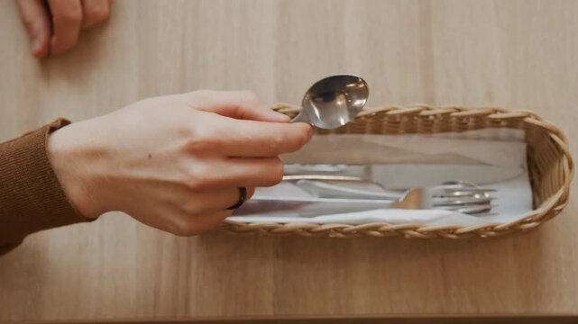Close up of hand reaching into frame to pick up silverware from wicker basket on wooden table. Hand grasps spoon then knife and fork sequentially while other utensils remain in woven