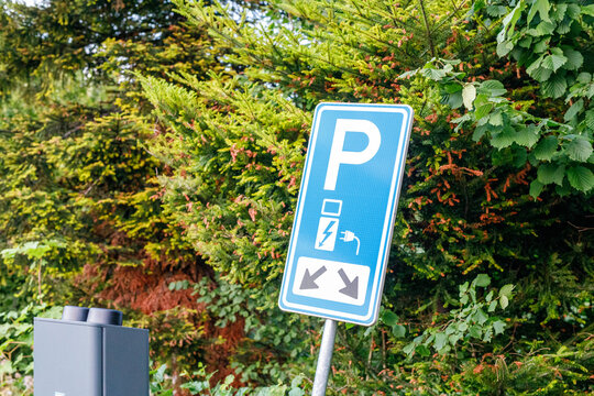 Blue sign indicating electric vehicle charging parking spots with a charging unit and green foliage.