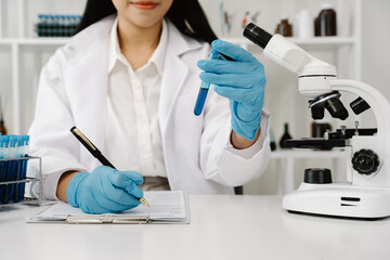 Fototapeta premium Scientific Discovery: A researcher in a lab coat and gloves meticulously records findings while holding a test tube filled with blue liquid, a microscope and various lab equipment in the background.