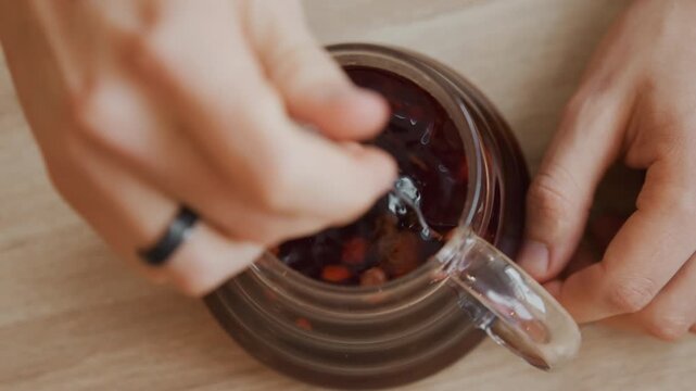 Close up of hands stirring dark liquid with spoon in glass pitcher on wooden surface. Hands hold and stir beverage creating gentle ripples one hand wears black ring. Natural light