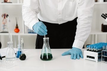 Fototapeta premium Scientist Mixing Liquid in Flask: A scientist in a lab coat and blue gloves carefully mixes green liquid in a flask, surrounded by laboratory equipment and test tubes.