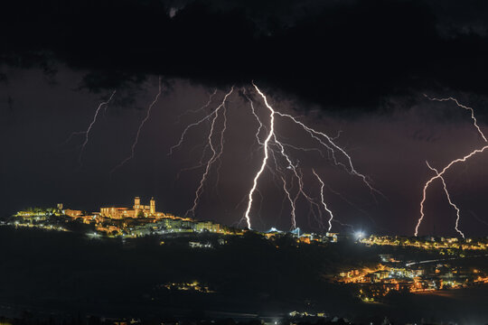 View of lightning strikes illuminating the night sky over the historic hilltop town of Montecosaro with glowing streetlights and dark storm clouds, Montecosaro, Marche, Italy.