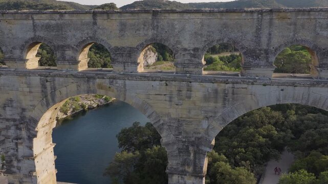 Close aerial through Pont du Gard arch over river forest path and tourists below