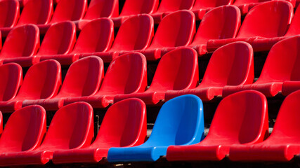 Fototapeta premium Blue seat standing out among rows of red plastic chairs at sports stadium, empty arena seating and contrast concept Photo
