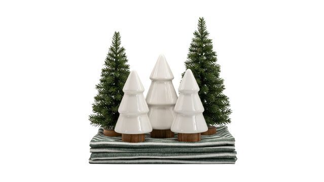 Three white ceramic christmas tree figurines with two artificial pine trees on a folded green striped fabric isolated on a transparent background