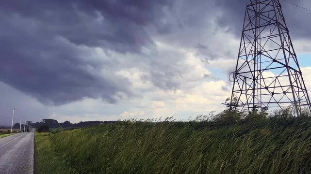 Low angle view of storm clouds and wind over a field  by a rural road with a high voltage transmission tower to the side in the midwest USA 