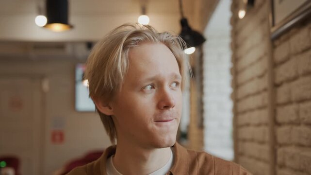 Close up of young man drinking from pink mug in indoor setting with soft lighting. subject is wearing brown shirt and background features blurred interior elements like hanging lights