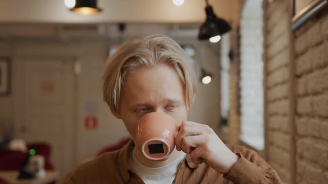 Close up of young man with short blonde hair wearing brown jacket and white shirt in indoor setting. Man holds cup near his mouth then gives thumbs up gesture while looking directly