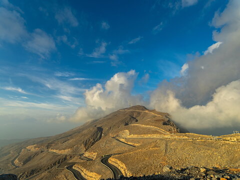 View of the winding mountain road leading up to Jebel Jais, surrounded by rugged rocky terrain and dramatic clouds under a blue sky in Ras Al-Khaimah, Ras Al Khaimah, United Arab Emirates.