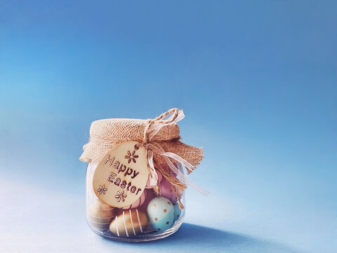 Glass jar filled with decorative Easter eggs on a blue background, burlap and ribbon accents, symbolizing holiday wishes