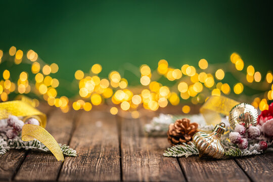 A Christmas background featuring golden ribbon, a pinecone, and shimmering ornaments framing wide rustic wood, set against green with twinkling lights and generous open copy space.