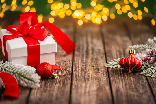 A Christmas background featuring a white gift box tied with red ribbon beside a pinecone, red ornaments, and frosted berries on rustic wood, lit by warm twinkling lights above with copy space.