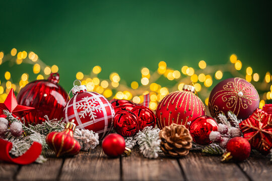 A Christmas background featuring an abundant arrangement of red baubles, a glittering star, and a pinecone nestled in frosted pine on rustic wood, set against green with twinkling lights.