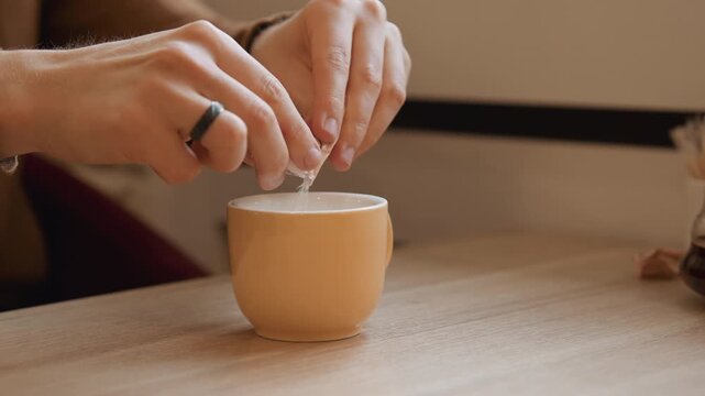 Close up of person hands tearing open and pouring sugar into ceramic cup on wooden table. scene features soft lighting with focus remaining steady throughout as details like texture detail
