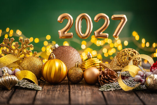 A Christmas background featuring luxurious gold 2027 balloon numbers surrounded by golden baubles, a snowflake, ribbon, and a pinecone on rustic wood, set against green with twinkling lights.