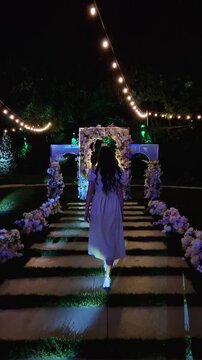 Woman walks down a flower-adorned aisle at night toward an illuminated wedding arch and chandelier. String lights overhead create a romantic evening ceremony atmosphere. Luxury event celebration