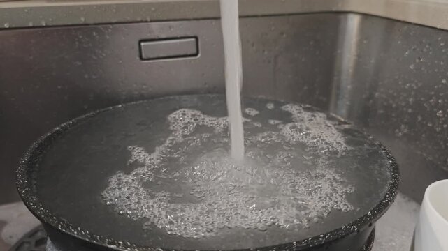 Close up of water running from a faucet, filling a black pan in a metal kitchen sink. Soapy bubbles form as the pan fills with water