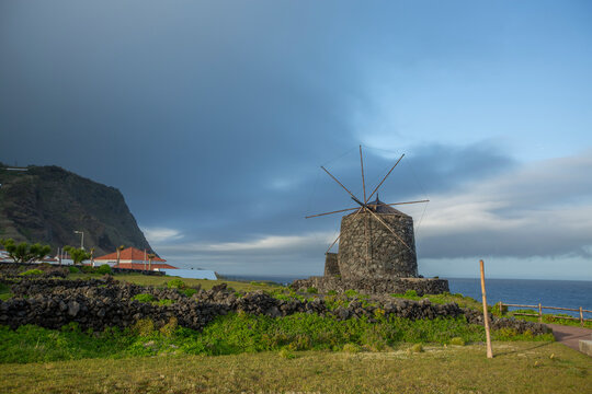 Corvo, Azores, Portugal Ancient stone windmill stands alone on rugged coastal cliffside. Dark storm clouds gather above, contrasting with bright blue sea. Lush green grass and wild vegetation surround