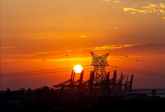 View of an electricity pylon and power lines silhouetted against a vibrant sunset sky with industrial port cranes in the background in Dubai, Dubai, United Arab Emirates.