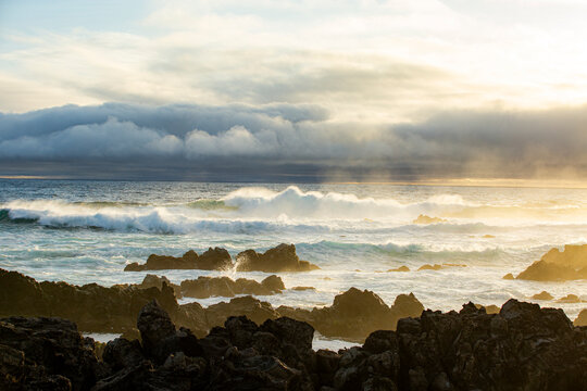 Corvo, Azores, Portugal Ocean waves crash against dark volcanic rocks at sunrise. Low clouds hover above the sea, catching golden light. Mist rises from the water, blending sky and sea. Dramatic light