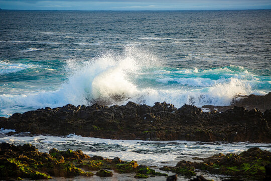 Corvo, Azores, Portugal Ocean waves crash against dark volcanic rocks. White foam bursts as water splashes upward. Green seaweed clings to wet stones near shore. Sky reflects gray-blue tones above chu