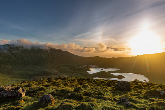 Corvo, Azores, Portugal Sunset bathes Corvo Island's lush green hills in golden light. Serene lake mirrors the sky's warm glow. Rolling clouds drift above rugged, moss-covered mountains. The scene fee