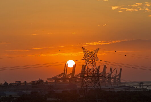 View of a high-voltage power pylon silhouetted against a vibrant orange sunset with industrial cranes in the background in Dubai, Dubai, United Arab Emirates.