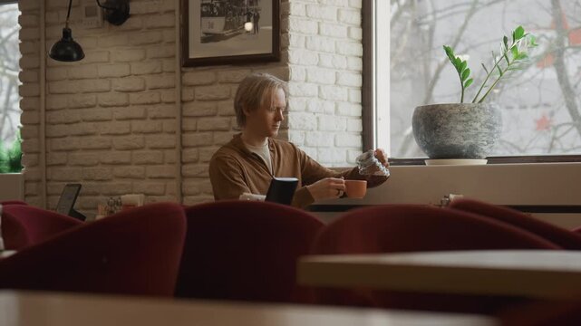 Close up of man sitting in empty cafe holding and pouring coffee into cup while looking out window. setting features white brick walls red chairs natural light from large windows framed