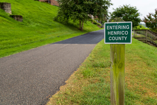 View of the Entering Henrico County sign on a wooden post beside a paved path and grassy hill in Henrico, Virginia, United States.