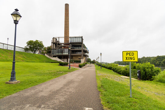 View of the modern industrial building with a tall brick chimney at Rocketts Landing, featuring a paved path and pedestrian crossing sign in Henrico, Virginia, United States.