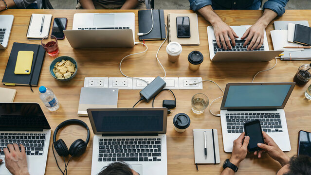 Overhead view of diverse professionals collaborating around a large wooden table filled with laptops and technology devices